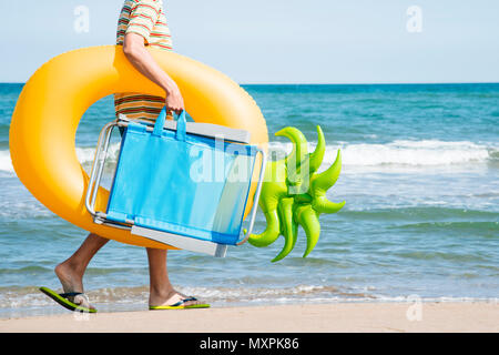 Primo piano di un giovane uomo caucasico la riva del mare, portante una sdraio sulla spiaggia e di un anello di nuotare in forma di ananas, andando o tornando dall'essere Foto Stock