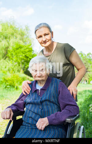 Donna anziana seduta in carrozzella con persone di mezza età figlia trascorrere del tempo di qualità per esterno Foto Stock