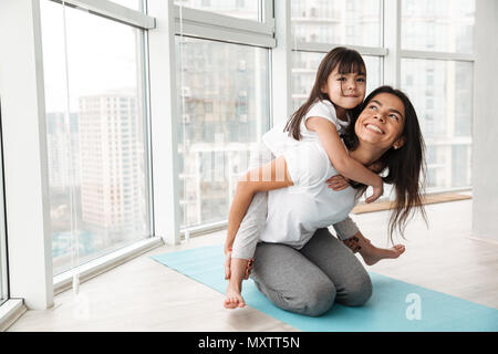 Ritratto di famiglia stupenda madre e bambino divertendosi e dando piggyback mentre fa sport esercita sul materassino yoga a casa Foto Stock