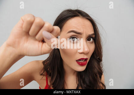 Chiudere l immagine di orinato brunette donna in abiti casual mostrando il suo pugno e guardando la telecamera su uno sfondo grigio Foto Stock