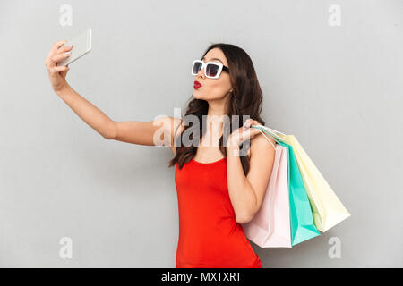 Giocoso brunette donna in abiti casual e occhiali da sole rendendo selfie sullo smartphone mentre si tiene dei pacchetti su uno sfondo grigio Foto Stock