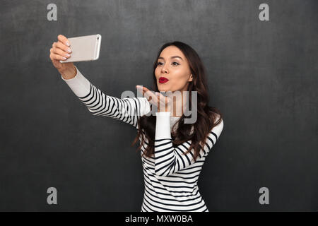 Il piacere bella bruna donna in maglione rendendo selfie sullo smartphone e invia aria kiss su sfondo nero Foto Stock