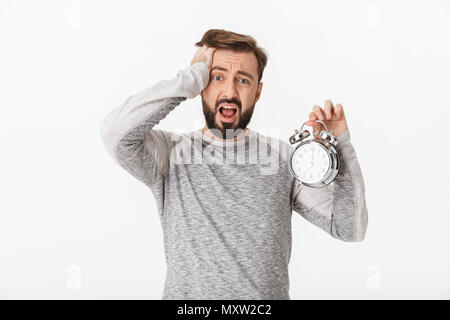 Foto di urlando spaventata giovane uomo isolato oltre il muro bianco sullo sfondo tenendo sveglia. Foto Stock