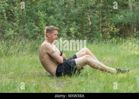 Barechested metà adulto 40s uomo caucasico facendo sit ups sul campo in erba all'aperto Foto Stock