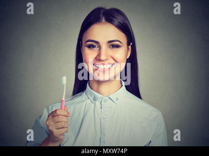 Luminose brunette holding spazzolino da denti e sorridente con denti bianchi guardando la fotocamera su grigio Foto Stock