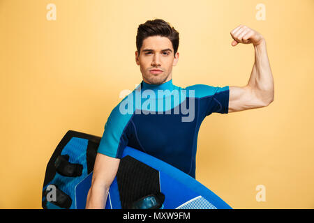 Ritratto di un fiducioso giovane uomo vestito in costume da bagno la flessione di muscoli mantenendo una tavola da surf isolate su sfondo giallo Foto Stock