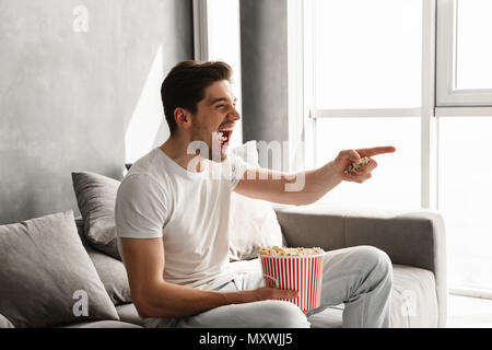 Felice estatica uomo seduto sul divano nel soggiorno e gesticolando dito sul televisore mentre mangia i pop corn Foto Stock