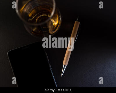 Whisky di vetro con una penna di legno e lo smartphone dal di sopra, superficie scura Foto Stock