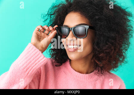 Immagine multicolore di donna americana 20s con acconciatura afro sorridente e vestita di nero moderni occhiali da sole isolate su sfondo blu Foto Stock