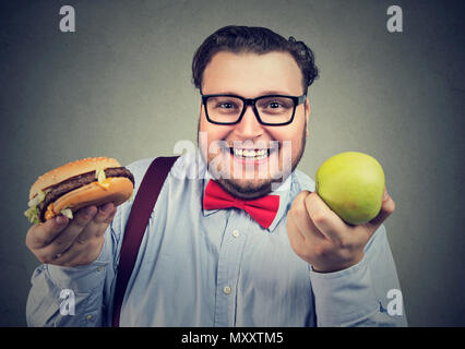 Felice giovane uomo chunky azienda verde mela fresca e hamburger rendendo la scelta e sorridente in telecamera Foto Stock