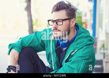 Tanga alla moda uomo in cuffie e giacca verde seduto sulla strada e guardando lontano nella contemplazione Foto Stock