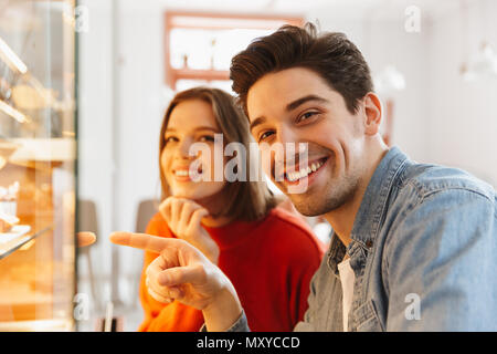 Bella giovane donna e uomo in abbigliamento casual sorridente e puntare il dito su gustosa dolci nel panificio accogliente Foto Stock