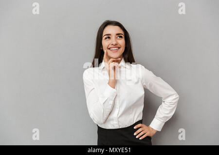 Ritratto di donna di successo 20s in maglia bianca cerca riposo e tenendo il dito a guancia mentre sorridente isolate su uno sfondo grigio Foto Stock