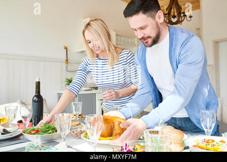 Ritratto della moderna coppia giovane impostazione tavolo per la cena mentre attende gli ospiti nella soleggiata sala da pranzo a casa, concentrarsi sulla coppia barbuto in primo piano Foto Stock