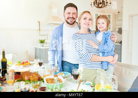 Ritratto di felice famiglia giovane con graziosi bambina in piedi alla cena di gala tabella con piatti deliziosi e sorridente alla fotocamera in moderno appartamento Foto Stock