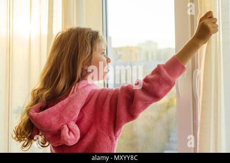Buona mattina. Bambine in pigiama apre le tende e guarda fuori dalla finestra Foto Stock