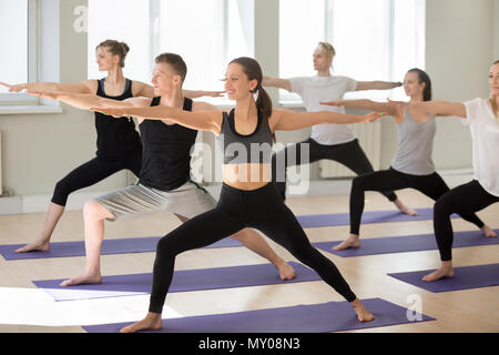 Un gruppo di giovani sportivi praticanti lezione di yoga, guerriero facendo due esercizi, Virabhadrasana 2 pongono, , yogi gli studenti che lavorano fuori dentro in sport c Foto Stock