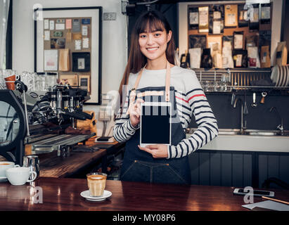 Donna asiatica barista indossare jean grembiule tenendo premuto lo schermo è vuoto compressa al cliente al banco bar con sorriso emozione,Cafe Restaurant service concetto,proprietario sm Foto Stock