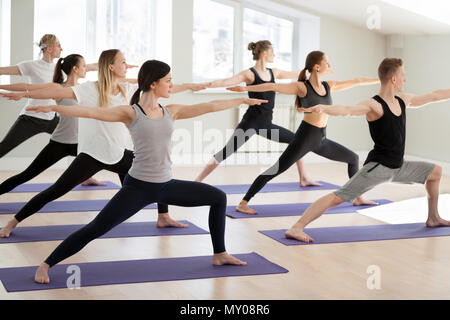 Un gruppo di giovani sportivi la pratica dello yoga, facendo del Guerriero II ESERCIZIO, Virabhadrasana 2 pongono, interni a lunghezza piena, yogi gli studenti che lavorano in sport Foto Stock