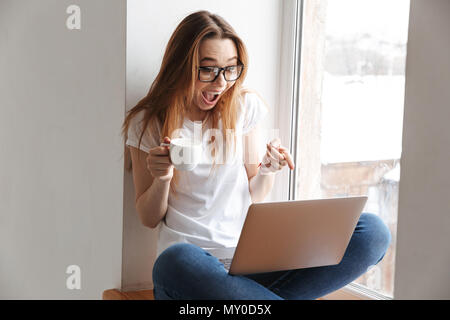 Scioccato donna felice in maglietta e occhiali seduto sul davanzale con tazza di caffè mentre si utilizza e puntando al computer portatile Foto Stock