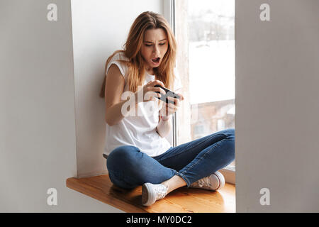 Giocoso donna in t-shirt seduta sul davanzale e la riproduzione tramite smartphone a casa Foto Stock