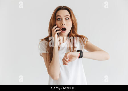 Scioccato la donna in abiti casual con orologio da polso parlando da smartphone e guardando la telecamera su uno sfondo grigio Foto Stock