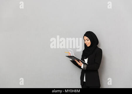 Ritratto di un sorridente giovane donna araba il lavoro con documenti isolate su uno sfondo grigio Foto Stock