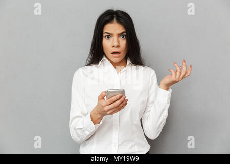 Foto di scioccato o indignato donna 30s in abbigliamento formale alla ricerca sulla telecamera tenendo lo smartphone nel canto isolato su sfondo grigio Foto Stock