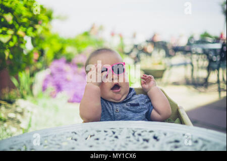 Un bambino è seduta al tavolo fuori e sta mettendo su un paio di occhiali da sole Foto Stock