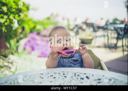 Un bambino è seduta al tavolo fuori e sta mettendo su un paio di occhiali da sole Foto Stock