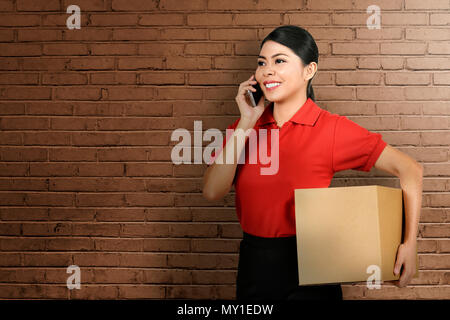 Bella asiatica donna consegna utilizzando il cellulare mentre portare il pacchetto con lo sfondo della parete Foto Stock