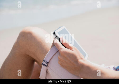 Primo piano di un giovane uomo caucasico sulla spiaggia, indossare pantaloncini rosa, utilizzando una tavoletta digitale Foto Stock