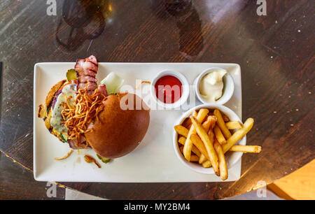 Tipico cibo da pub inglese: Cheeseburger in un panino con pancetta e patatine fritte (patatine) con ketchup di pomodoro e salse di maionese serviti su un piatto bianco Foto Stock