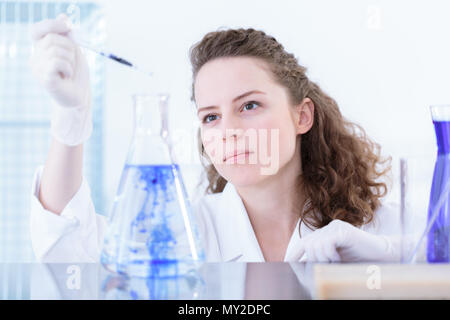 Farmacia in camice bianco tenendo una pipetta e aggiunta di una sostanza di colore blu di un pallone da laboratorio, rendendo una miscela Foto Stock