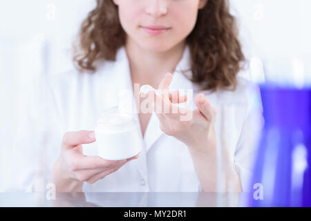 Farmacia femmina in camice da laboratorio tenendo un contenitore bianco e presentando la lozione sul suo dito Foto Stock