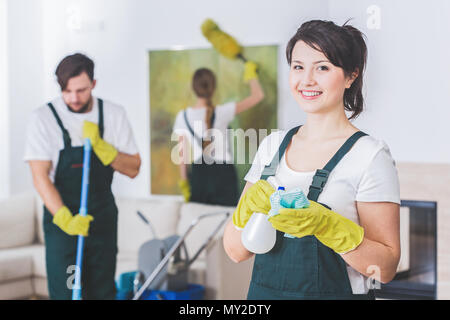 Gruppo di giovani e duro lavoro di pulitori professionali in appartamento sporco. Donna con soluzione detergente e panno contro sfondo sfocato Foto Stock