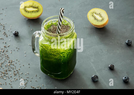 Verde, frullato sano in un vasetto di vetro con una cannuccia e kiwi, mirtilli e chia semi sparsi intorno Foto Stock