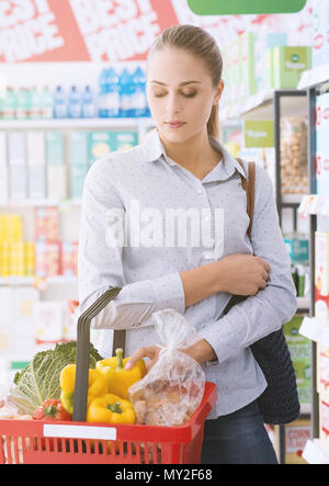 Giovane donna gli acquisti al supermercato, lei sta portando un cesto di generi alimentari riempito con verdure fresche Foto Stock