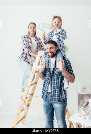 La famiglia felice rinnovando la loro nuova casa, sono in posa insieme con una scala e un rullo di vernice, il padre è piggybacking suo figlio Foto Stock