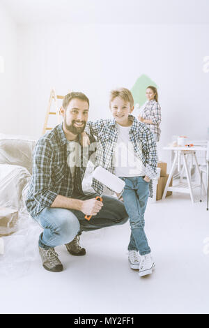 Home makeover, decorazione e verniciatura: un padre è in posa con il suo figlio e trattenimento di un rullo di vernice, sua moglie è la pittura pareti sullo sfondo Foto Stock