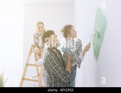 Felice famiglia giovane rinnovando la loro casa, il padre è in possesso di suo figlio ed egli lo aiuta a dipingere una parete con un rullo di vernice, la madre è stand Foto Stock