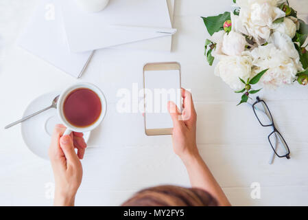 Area di lavoro con la ragazza mani smart phone con schermo vuoto e la tazza di tè, bicchieri, notebook, peonia bianco sul tavolo ligneo. Vista superiore del femminile scrivania in ufficio. Pianificazione. Freelance di luogo di lavoro. Foto Stock