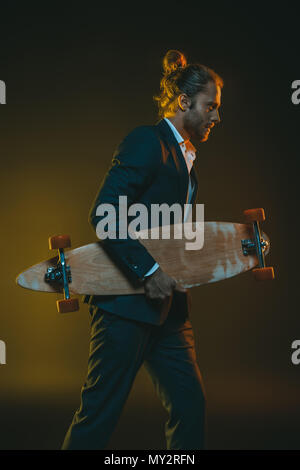 Vista laterale del giovane uomo caucasico in smoking in esecuzione con lo skateboard in mani Foto Stock