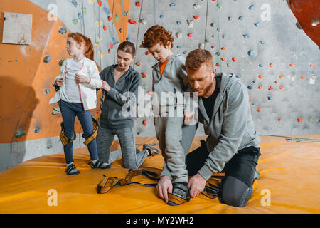 I genitori che fissano i loro figli al sistema di cavi per la scalata di una parete con impugnature in palestra Foto Stock