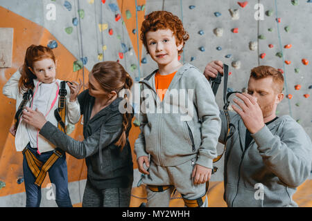 A mezza lunghezza shot dei genitori che fissano i loro figli al sistema di cavi per la scalata di una parete con impugnature in palestra Foto Stock