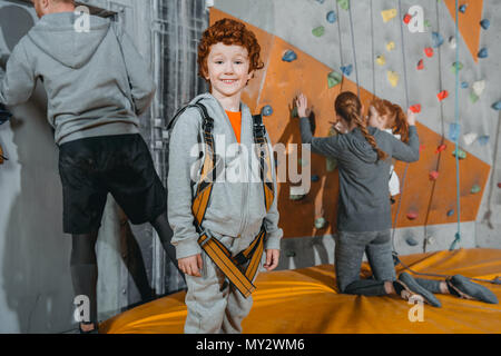 A mezza lunghezza shot di un po' di red-headed boy in un cablaggio di arrampicata in piedi in palestra e guardando la telecamera, con pareti di arrampicata in background Foto Stock