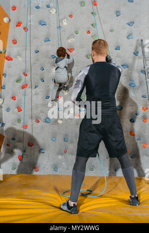 A tutta lunghezza shot di Little Boy salendo una parete con impugnature in palestra e suo padre tenendo il fissaggio corda Foto Stock