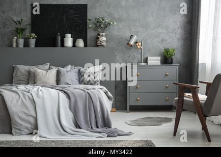 Cabinet grigio accanto al letto in camera da letto interno con poltrona e poster nero sulla parete. Foto reale Foto Stock