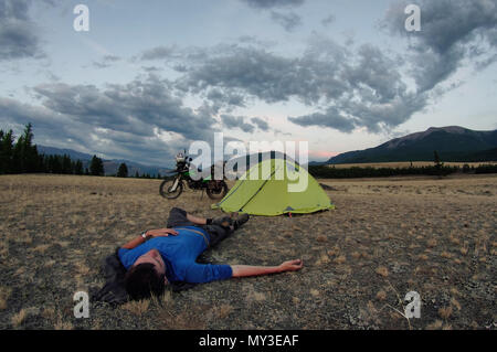 Uomo disteso sul terreno e camp tenda con moto enduro da soli viaggiatori steppa il paesaggio sullo sfondo di alte montagne al tramonto sulle montagne di Altai Foto Stock