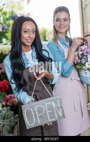 Sorridente multietnica di giovani donne in grembiuli tenendo aperte segno e fiori freschi in negozio di fiori Foto Stock